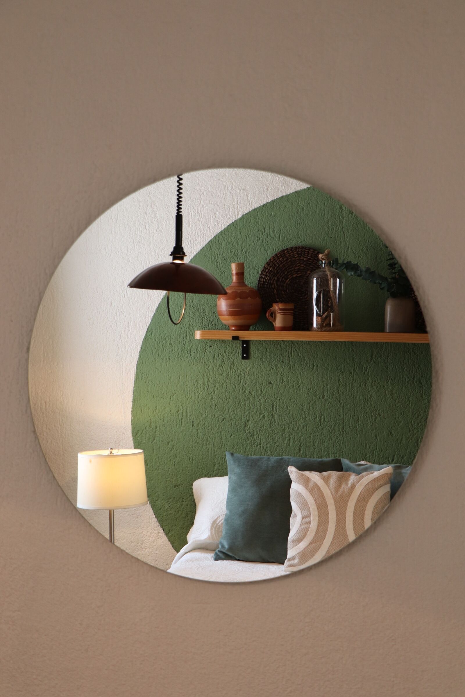 Uno specchio circolare che riflette una stanza con un muro verde, uno scaffale in legno con oggetti decorativi, una lampada e un letto con cuscini a fantasia.