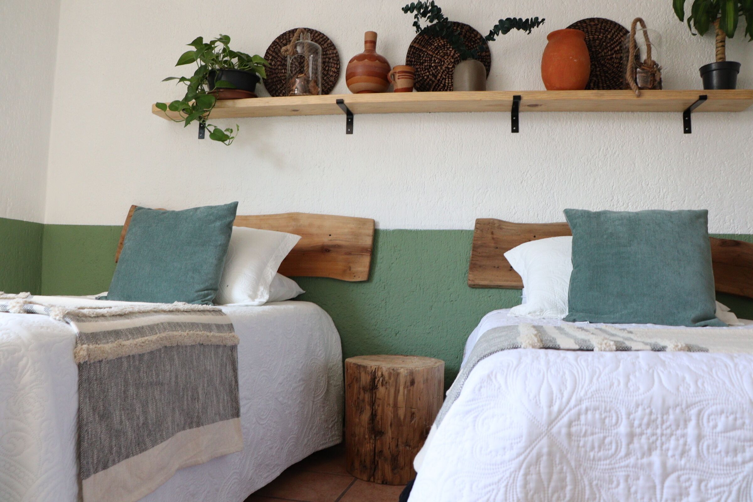 A bedroom with two beds, each with a wooden headboard, a bedside table, and a small stool. There is a shelf above the beds with decorative items.