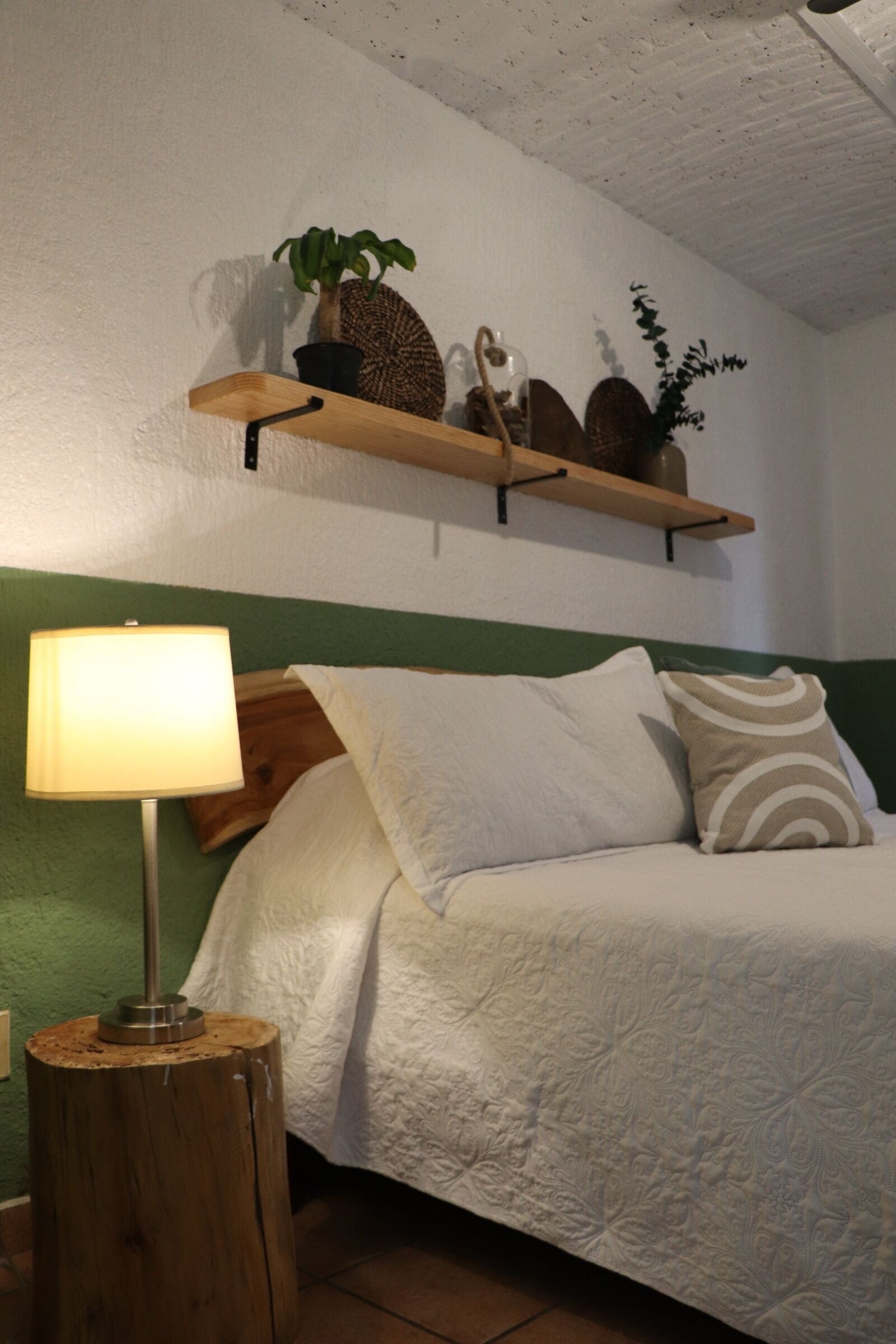 A bedroom with a bed, a bedside lamp, a wooden nightstand, a shelf with decorative items, and a green wall.