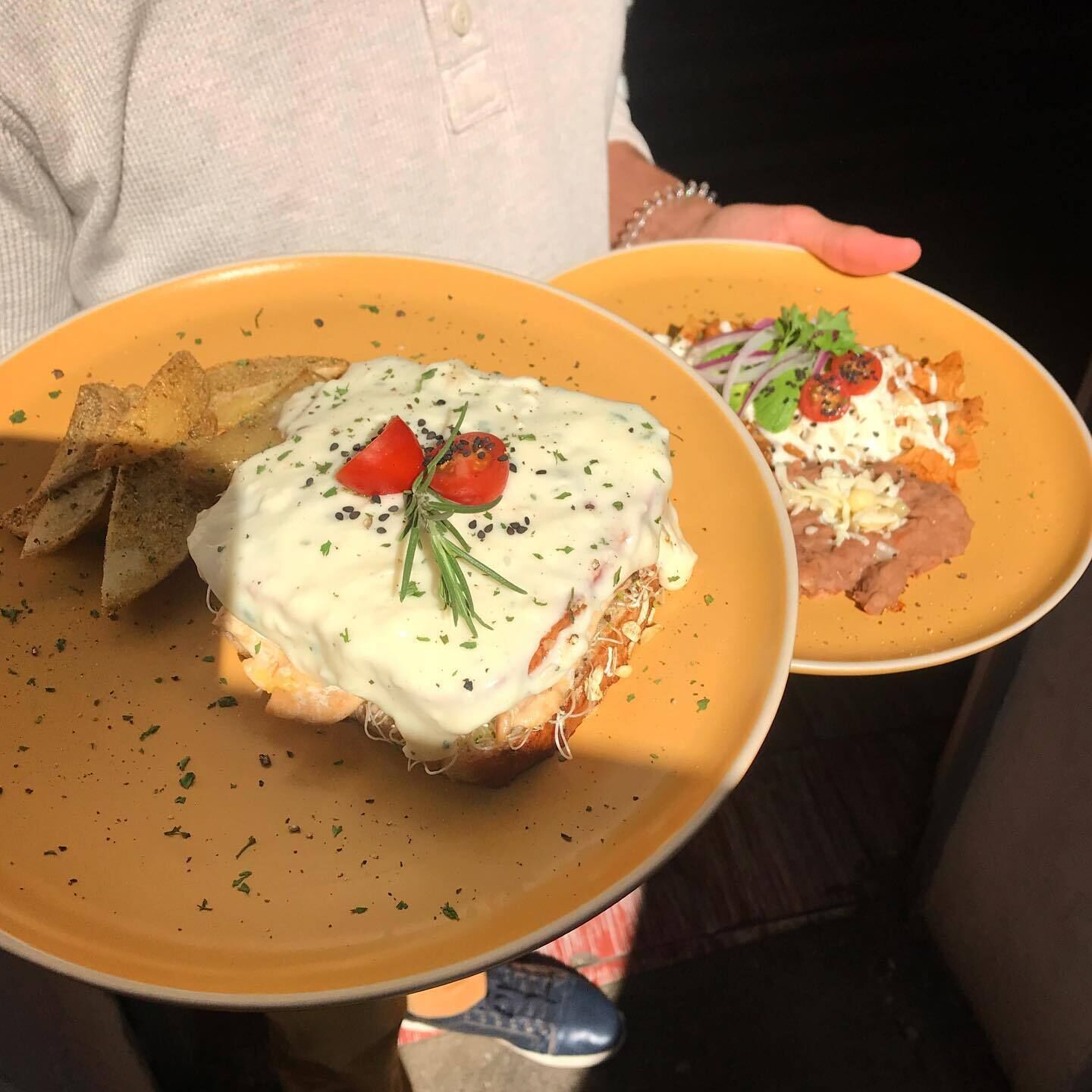 A person holding two plates with food, one with a white sauce and tomato garnish, and the other with beans and a green topping.