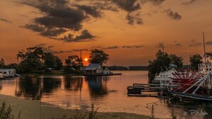 A sunset over a calm lake with boats docked, a small house, and a windmill. - Floating Cottage on the River "Nantucket" (Sabula)