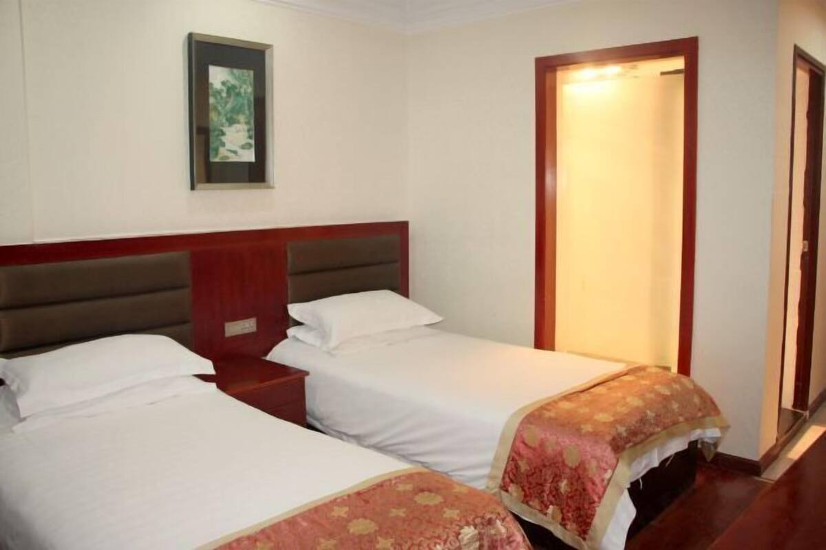 Een hotelkamer met twee enkele bedden, een houten hoofdbord, een ingelijst schilderij aan de muur en een deur die naar een andere kamer leidt.