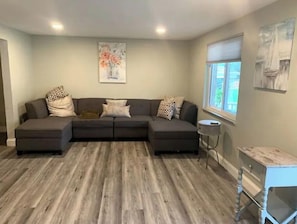 Un salon moderne avec un canapé d’angle, un plancher en bois, un tableau au mur et une fenêtre avec des stores.