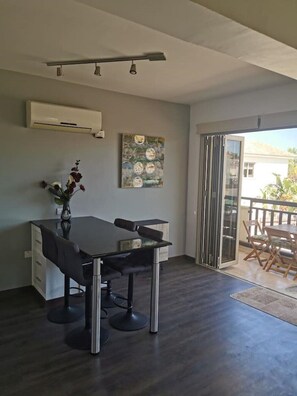 A modern dining area with a glass table, black chairs, and a vase of flowers. There is a ceiling light fixture and an air conditioning unit on the wall.
