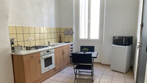 Een functionele keuken met een fornuis, oven, spoelbak en een kleine koelkast. Er is een eettafel met twee stoelen en een raam dat voor natuurlijk licht zorgt.