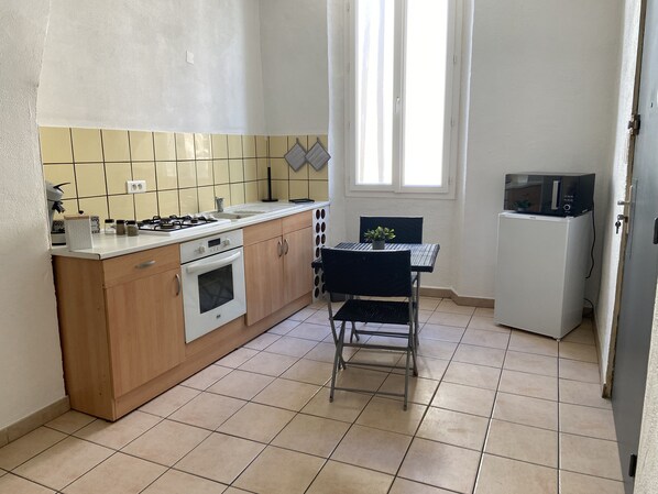 A compact kitchen with a stove, oven, sink, and a small refrigerator. There's a dining table with two chairs and a window providing natural light. - Studio in the center of Digne les bains (Digne les bains)
