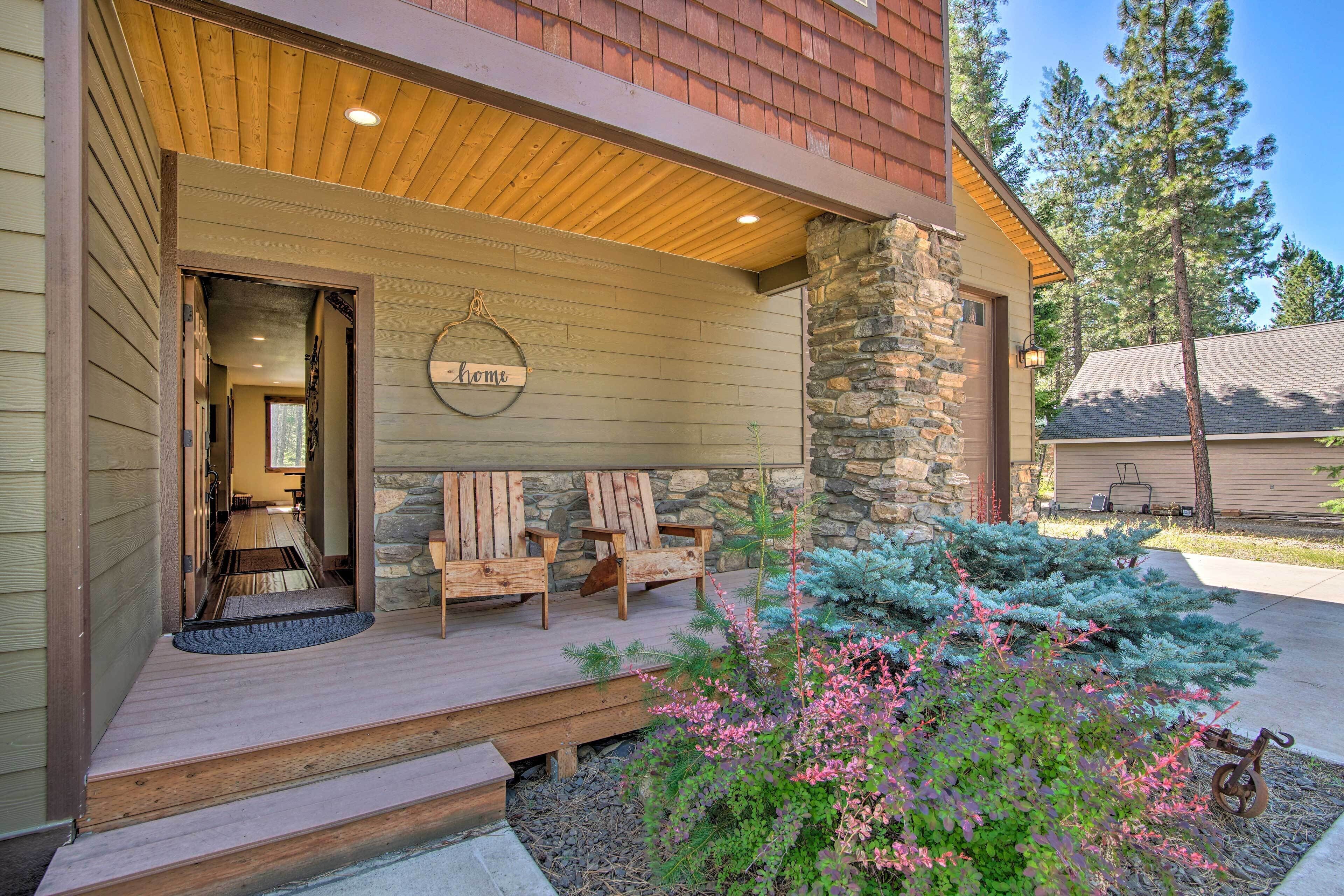 A wooden porch with two chairs and a welcome sign, a stone pillar, and a small garden with pink flowers.