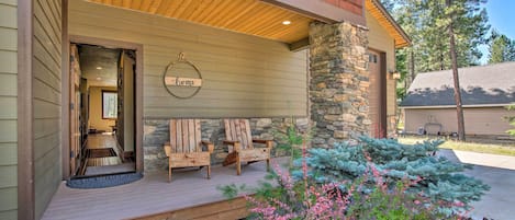 A wooden porch with two chairs and a welcome sign, a stone pillar, and a small garden with pink flowers.
