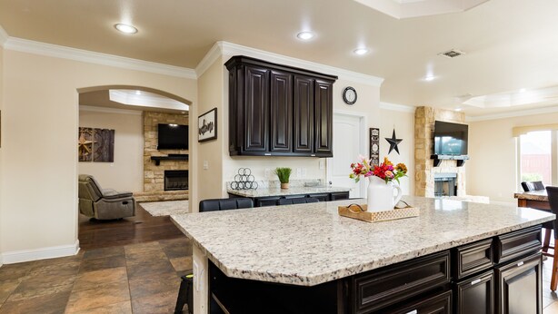 A spacious kitchen with dark cabinetry, a granite countertop, and a stone fireplace. - The ultimate resort ranch style pool home on nearly a half acre! (Lubbock)