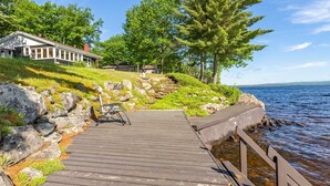 A wooden dock leading to a house with a porch, surrounded by greenery and overlooking a body of water.