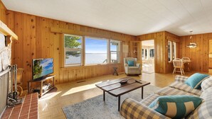 A wooden cabin with a large window overlooking a body of water, a television, a sofa, and a dining area.
