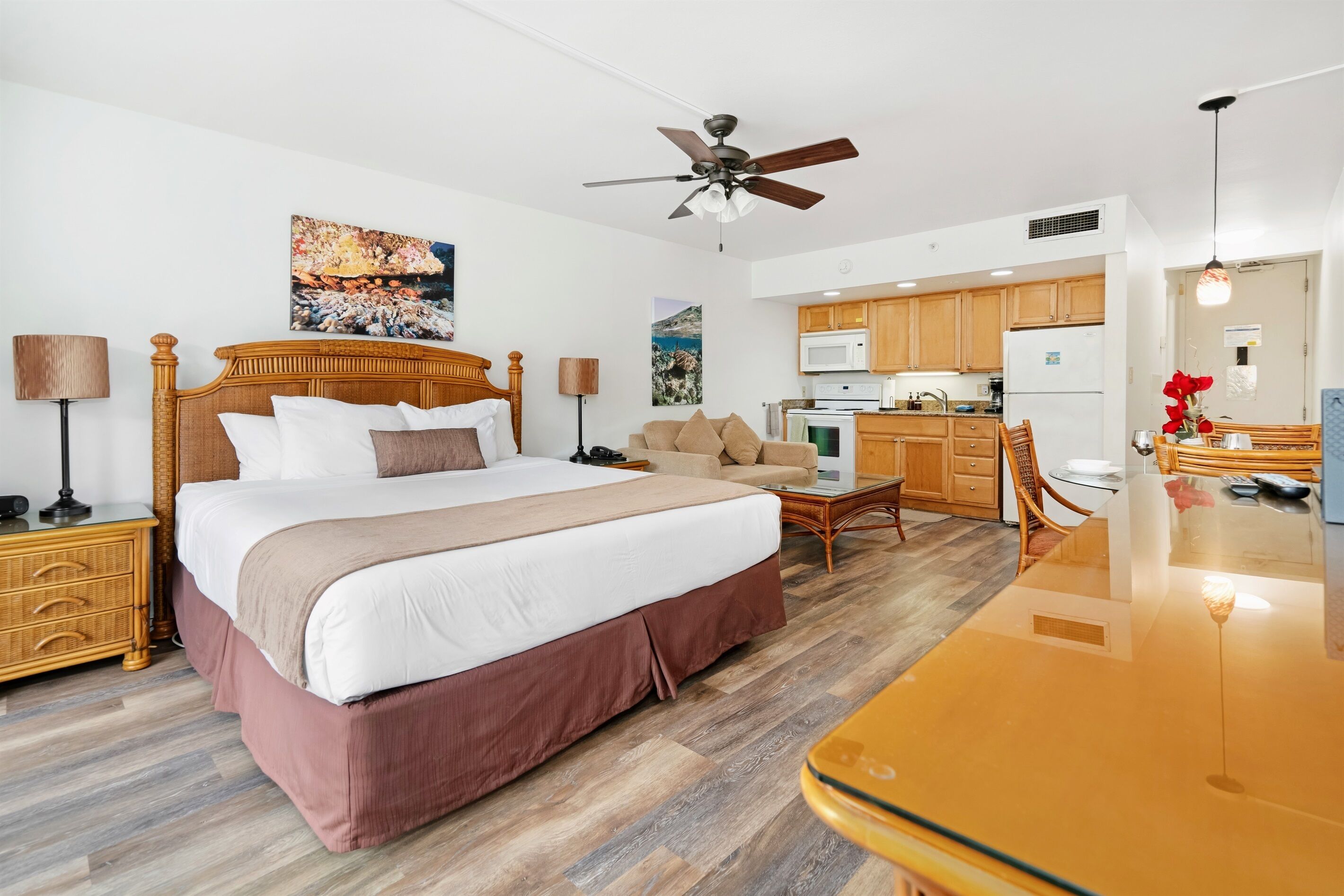 A hotel room with a large bed, a desk, a kitchenette, and a ceiling fan.