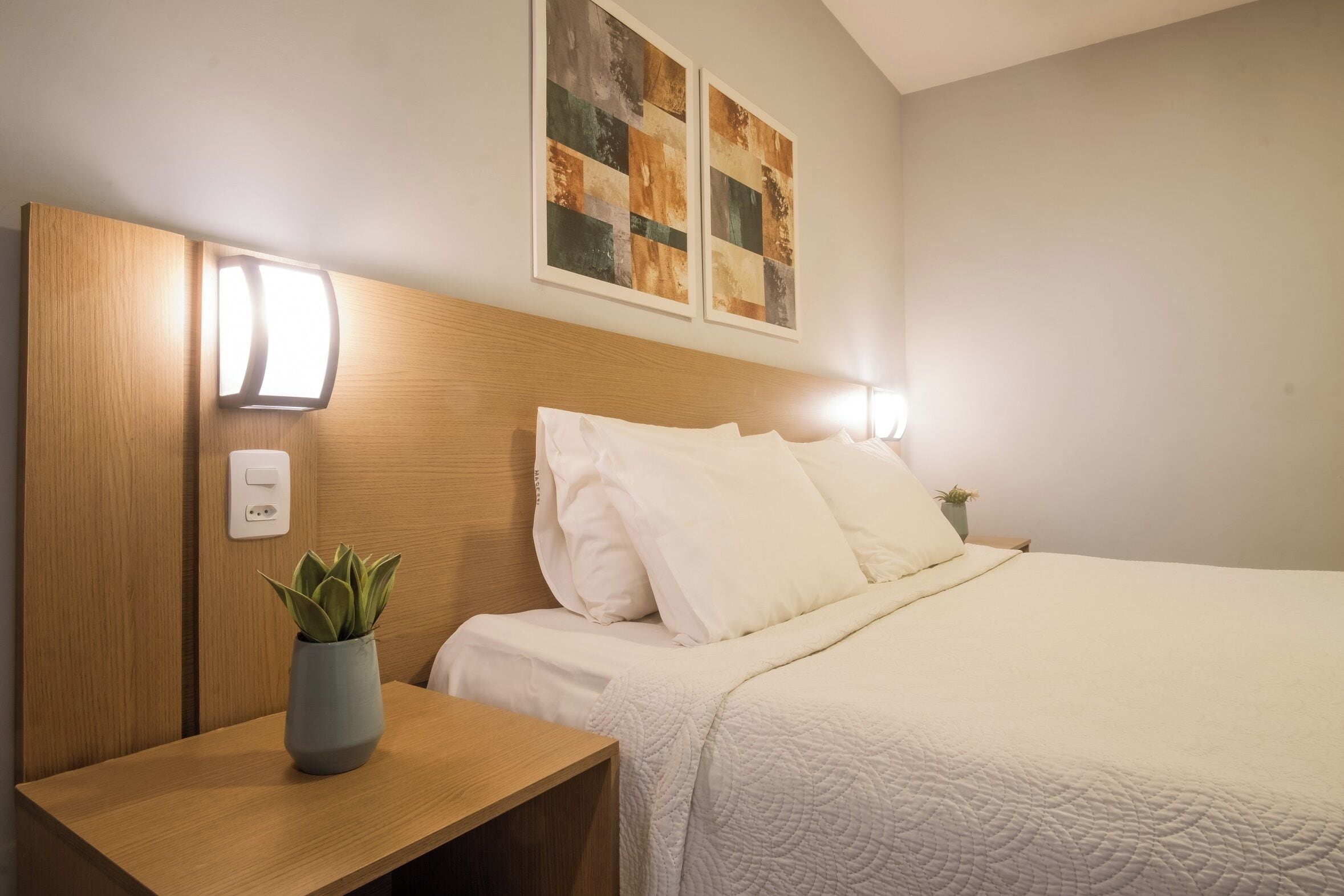 Una cama bien hecha con sábanas blancas, una mesita de noche con una planta y dos lámparas fijadas a la pared.