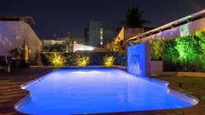 A swimming pool at night with surrounding greenery and buildings in the background. - Marezzi Hotel (Aracaju)