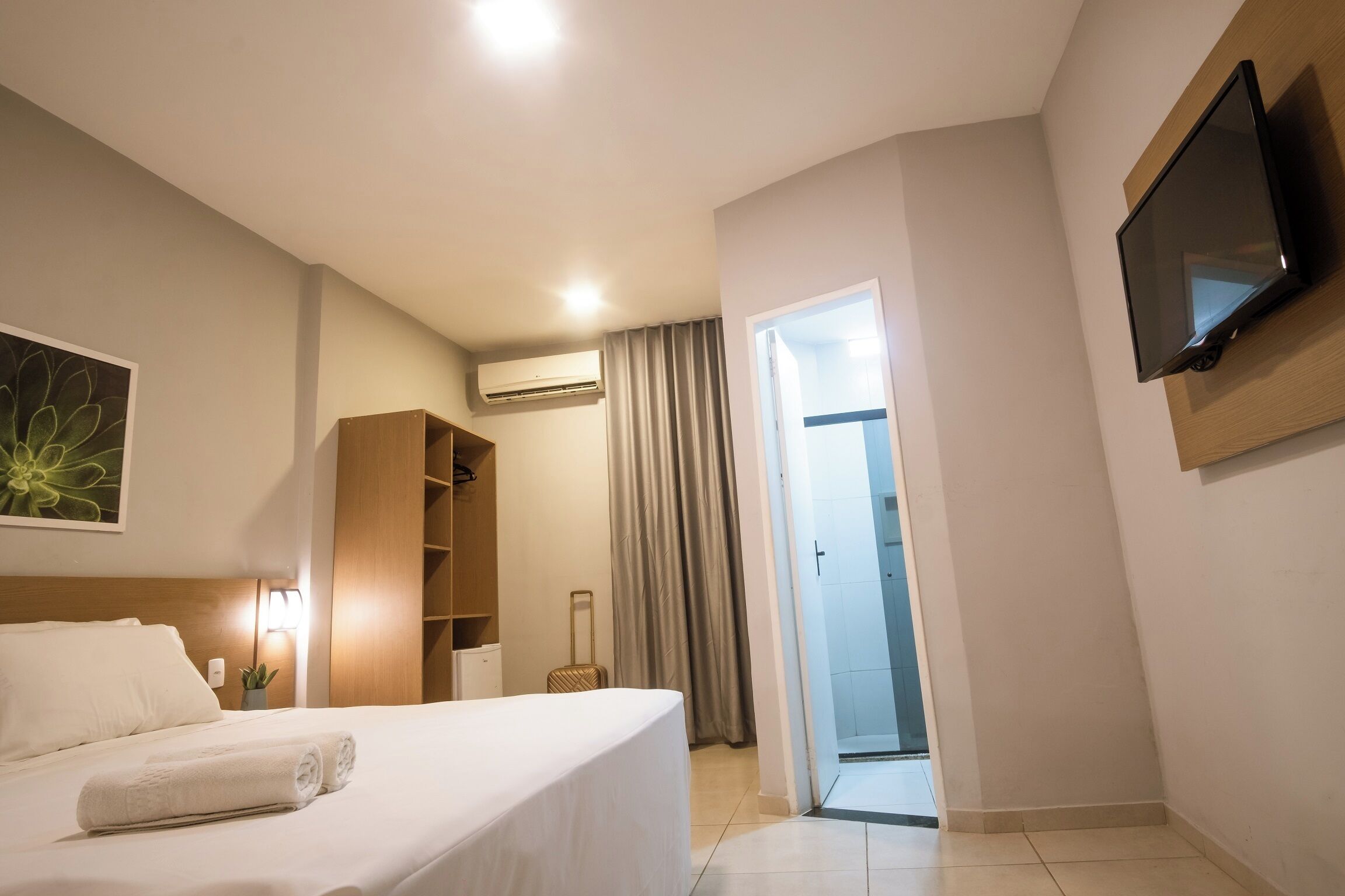 Quarto de hotel com cama, televisão presa à parede, guarda-roupa e porta deslizante para uma varanda.