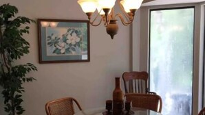 A dining area with a glass table surrounded by wicker chairs, a chandelier, a framed floral painting, and a large window.