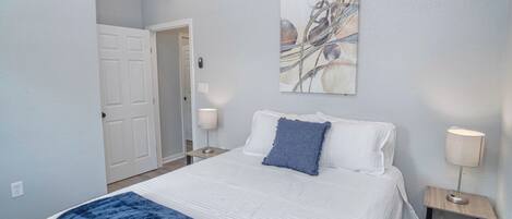 Une chambre à coucher avec un lit, un plaid bleu, une tête de lit blanche, une table de chevet avec une lampe et une porte donnant sur une autre pièce.