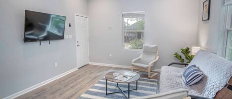 Un salon avec un téléviseur à écran plat fixé au mur, un plancher en bois, un tapis à rayures, un canapé, un fauteuil et une fenêtre avec des stores.