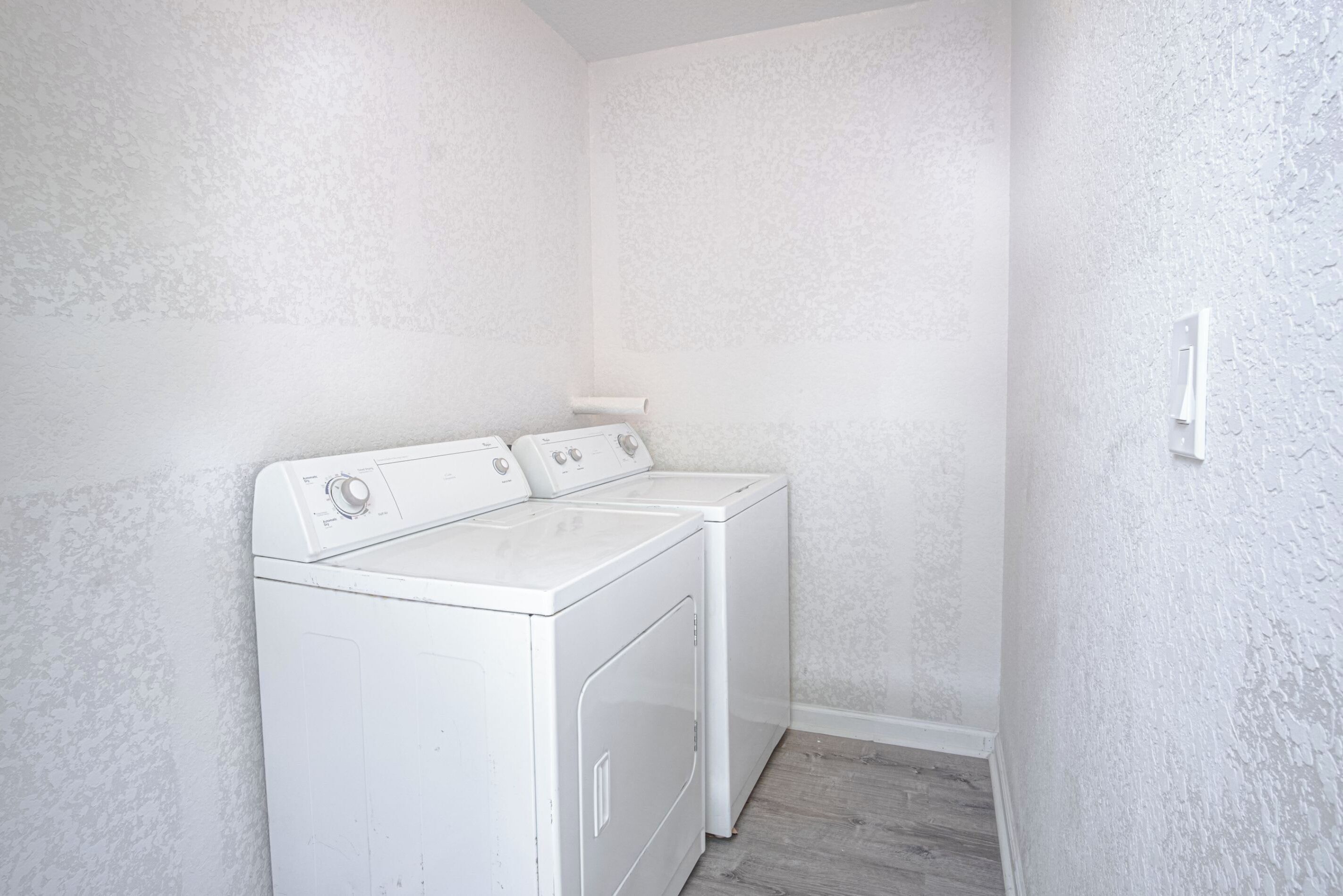 A laundry room with two white front-loading washers and dryers, a wooden floor, and textured white walls.