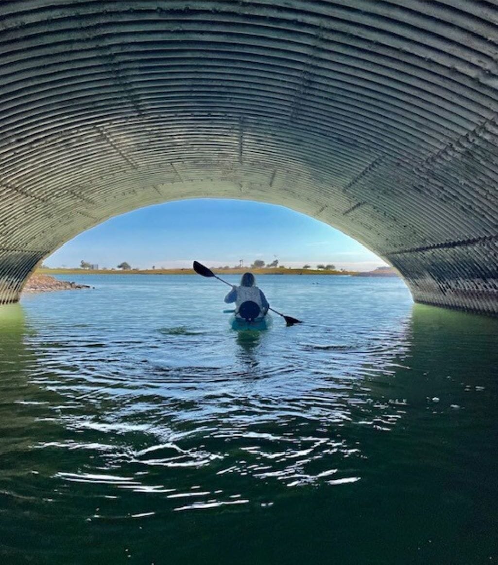 A kayaker under a bridge with calm water and a clear sky.