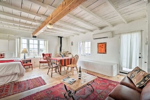 Een ruime kamer met houten plafond, een eethoek met tafel en stoelen, een bed met rode deken, en een groot raam.