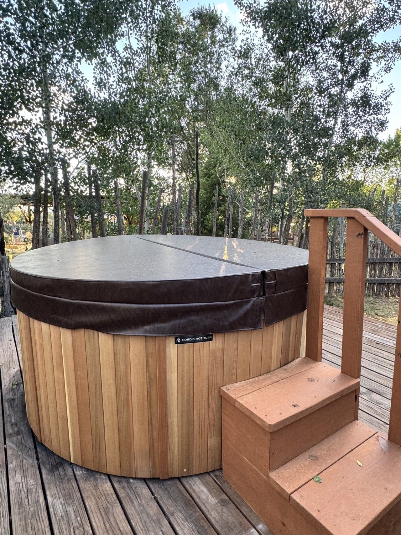 Ein Whirlpool mit Holzaußenverkleidung und schwarzem Deckel, gelegen auf einer Holzterrasse mit einer Holztreppe, die hinaufführt.