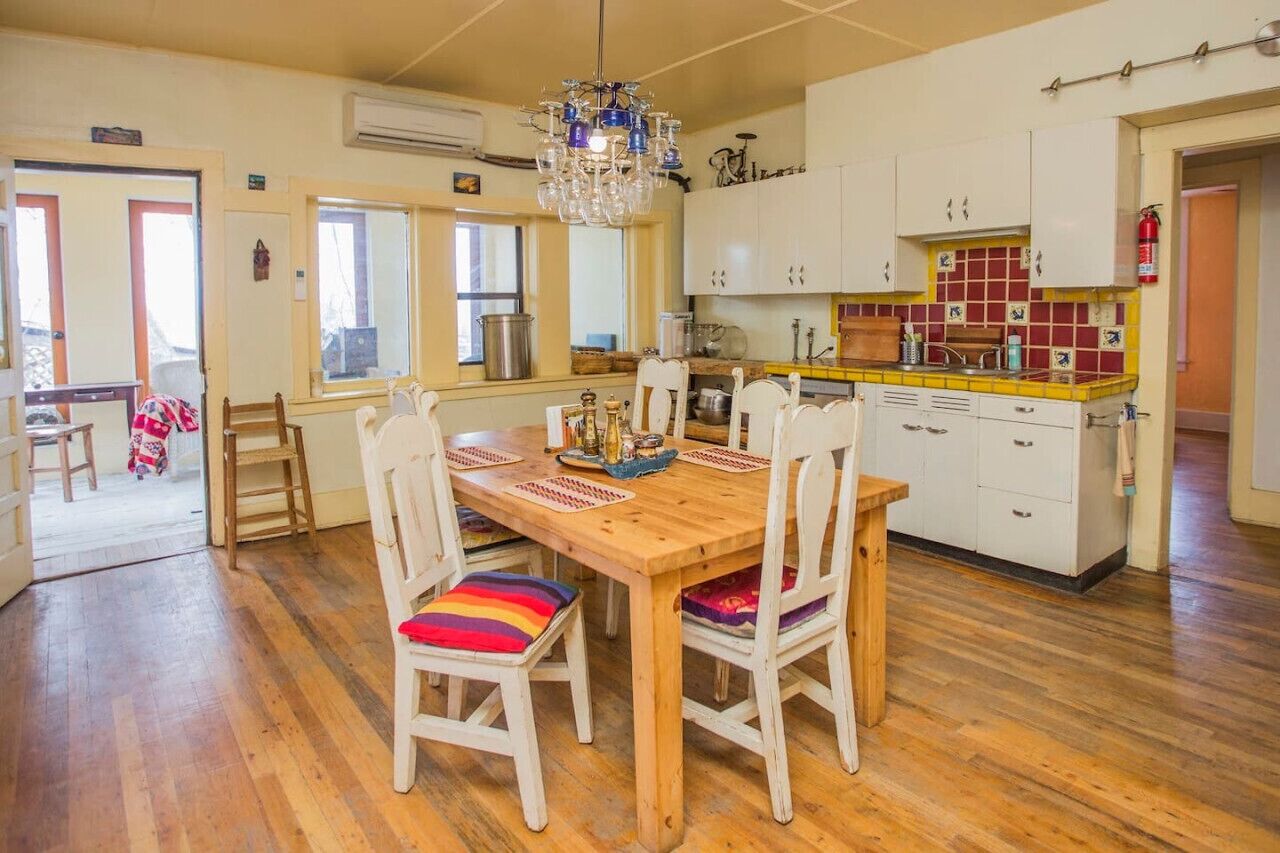A dining area with a wooden table and white chairs, a kitchen with white cabinets and a tiled backsplash, and a chandelier.