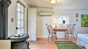 Una habitación con suelo de madera, una mesa con sillas, una cama, un ventanal con persianas y un aire acondicionado de pared.