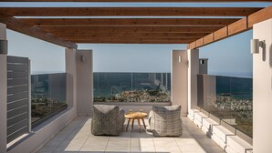 A rooftop terrace with a wooden pergola, two wicker chairs, a small round table, and a glass balustrade overlooking a coastal cityscape. - Villa Heora - The Floating Residence (Tria Monastiria)