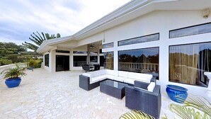Une terrasse extérieure moderne, avec un canapé blanc, des meubles en osier et de grandes fenêtres.