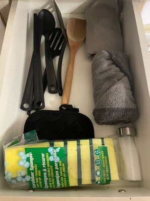 A drawer containing a black spatula, a wooden spoon, a gray towel, a black bag, and a package of scrubber sponges.