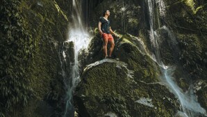 Eine Person steht auf einem mit Moos bewachsenen Felsen, im Hintergrund ein Wasserfall in bewaldeter Landschaft.
