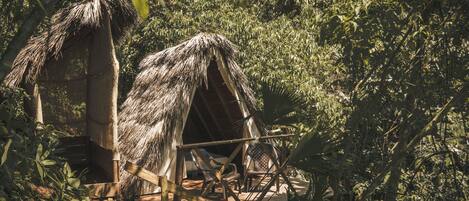 Una cabaña con techo de paja, terraza de madera y sillas, rodeada de follaje denso.
