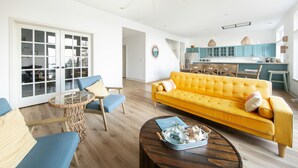 Un salon lumineux avec un canapé jaune, des fauteuils bleus, une table basse ronde et une cuisine en arrière-plan.