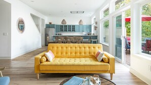Un salon lumineux avec un canapé jaune, une cuisine à l’arrière-plan et de grandes fenêtres.