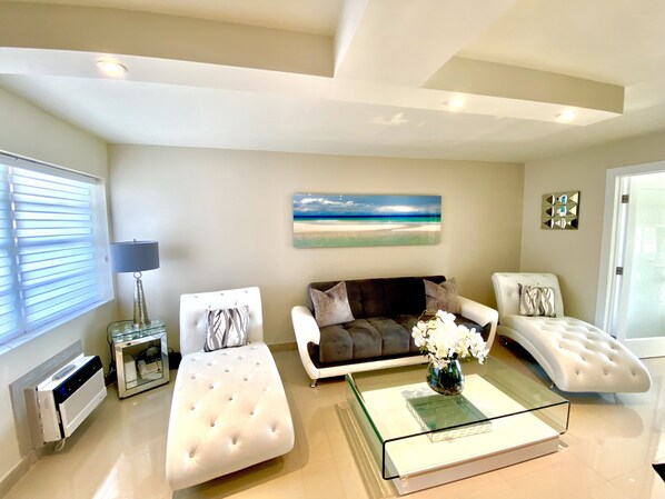 Un salón moderno con una mesa de centro de vidrio, un sofá, dos sillones, una lámpara y un cuadro con motivos playeros en la pared.