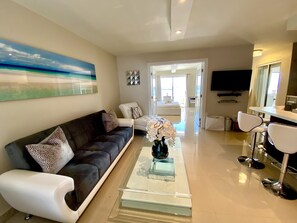 Una sala de estar moderna con un sofá oscuro, sillones blancos y una mesa de centro de vidrio con un jarrón de flores. Se ve un cuadro y una decoración mural geométrica.
