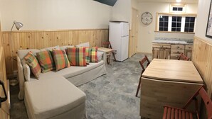Una habitación acogedora con sofá, una pequeña mesa, una silla, refrigerador y un reloj en la pared.