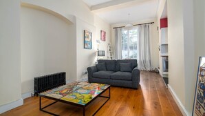 Un salón con un sofá gris, una mesa de centro con la portada de un cómic, un radiador y una ventana con cortinas.