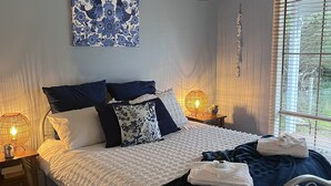 A bedroom with a bed, bedside lamps, a hanging light fixture, a window with blinds, and a wall-mounted artwork. - Cosy Pet Friendly Beach Cottage (Golden Beach)