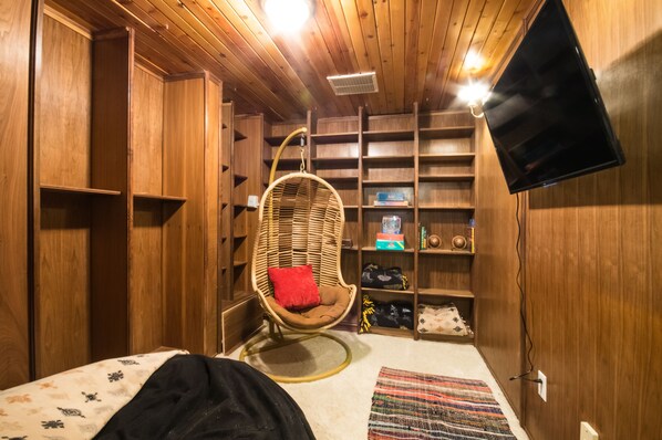 A wooden-paneled room with a bed, a wicker chair with a red cushion, a television on the wall, and shelves with various items. - 5 bd, 4 bth, A/C, new mattresses, hot tub, waterfalls, walk to town, trails! (Green Mountain Falls)