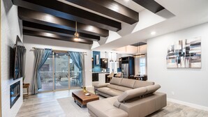 A modern living room with a large sectional sofa, a flat-screen TV mounted on the wall, and a dining area with a kitchen in the background. - VILLA WITH MASTER SUITE CLOSE TO SHOPPING 4 BR 3BA (EL PASO)
