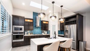 A modern kitchen with dark cabinetry, stainless steel appliances, a central island with bar stools, and pendant lighting. - VILLA WITH MASTER SUITE CLOSE TO SHOPPING 4 BR 3BA (EL PASO)