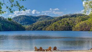A lakeside view with wooden Adirondack chairs and a table on the shore, surrounded by forested hills. - NEW High-Speed WiFi! Movie Theater, Pool Table & Hot Tub in Bear Lake Reserve!! (Tuckasegee)
