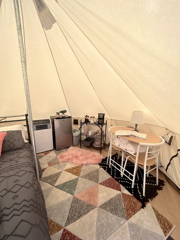 A tent with a bed, a table with a chair, a heater, a small shelf with items, and a patterned rug.