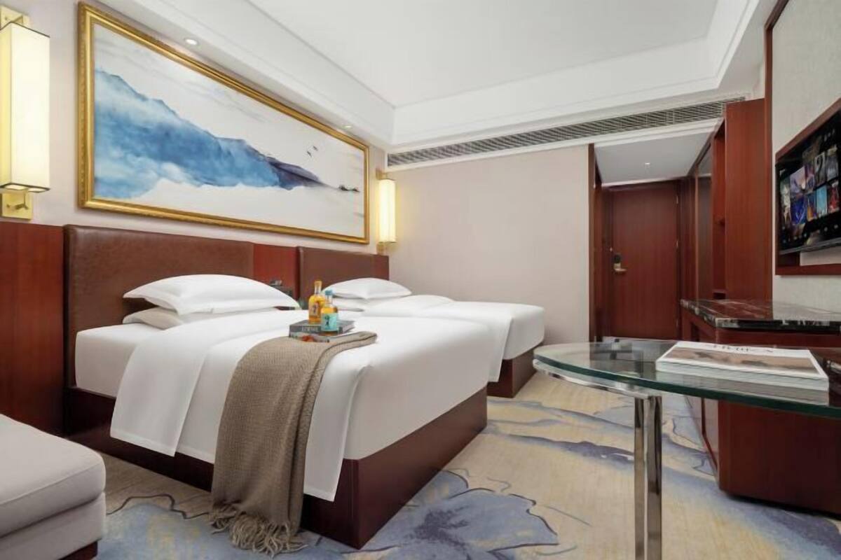 A hotel room with a bed, bedside tables, a glass coffee table, a painting on the wall, and a television.