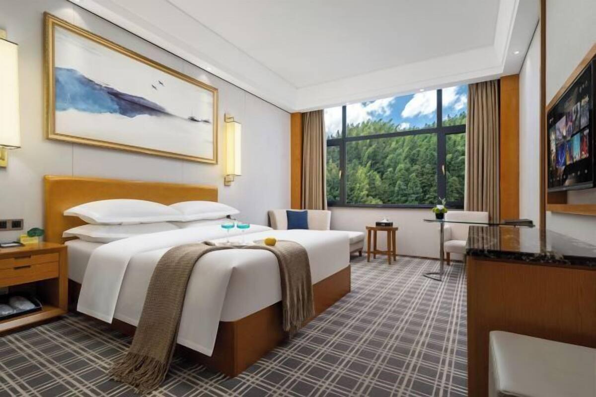 A hotel room with a large bed, a bedside table, a small table, a chair, a window with a view of trees, and a painting on the wall.