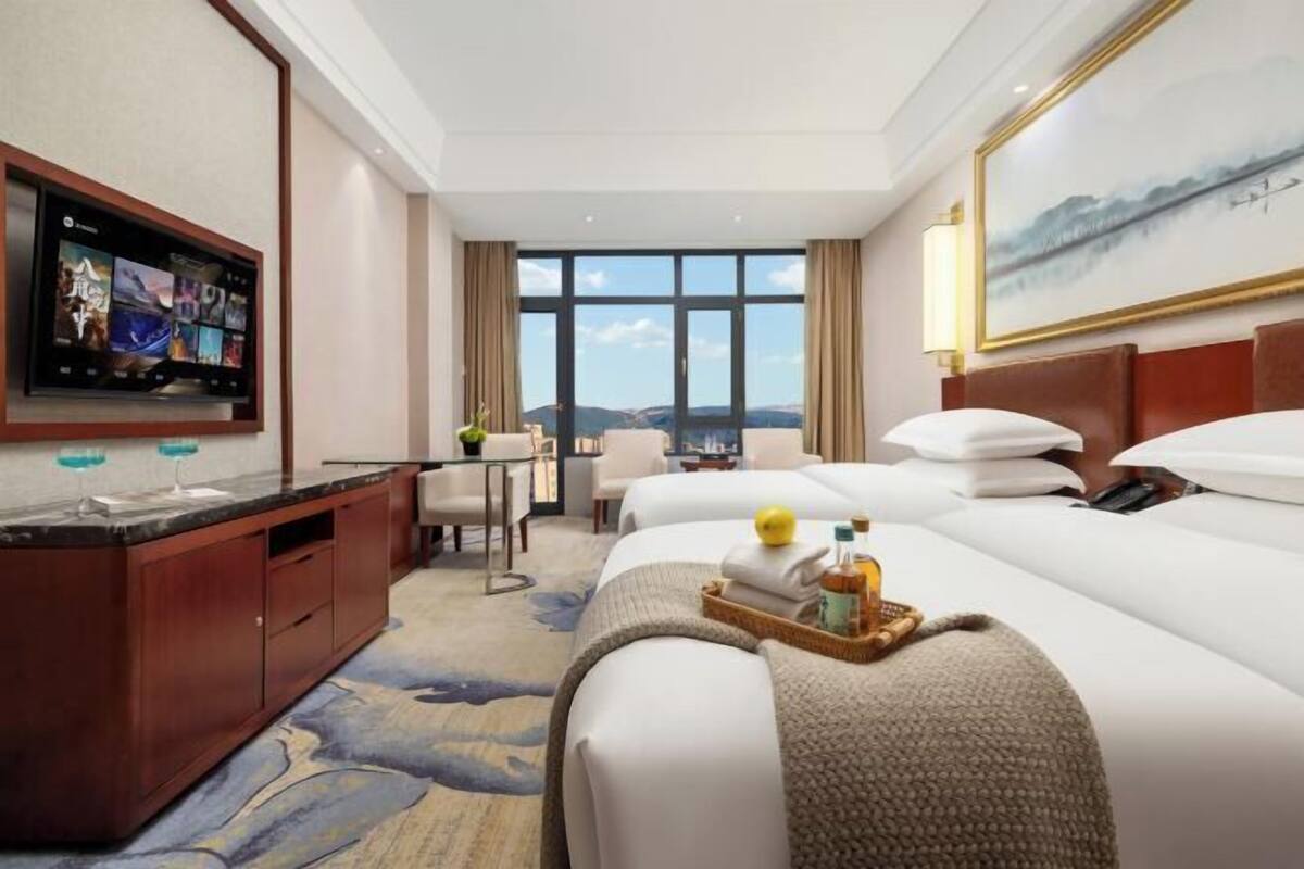 A hotel room with a large TV, a desk with a chair, two beds, and a view of the city.