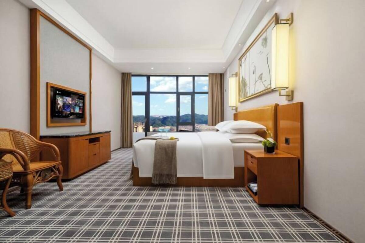 A hotel room with a bed, a television, a view of the mountains, and a large window.