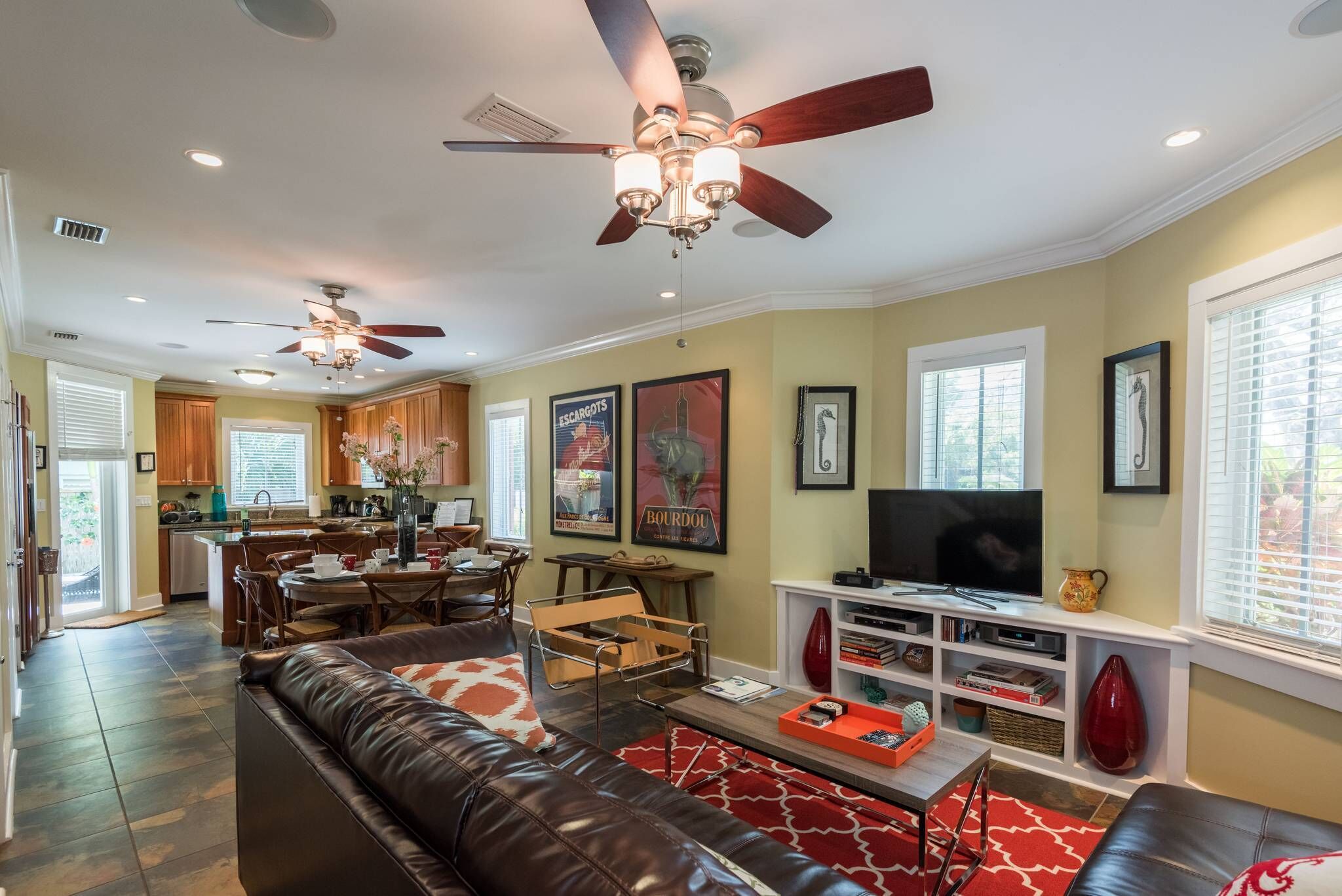 A spacious living room with a brown leather sofa, a coffee table, and a TV. There is a dining area with a table and chairs, and a kitchen in the background.
